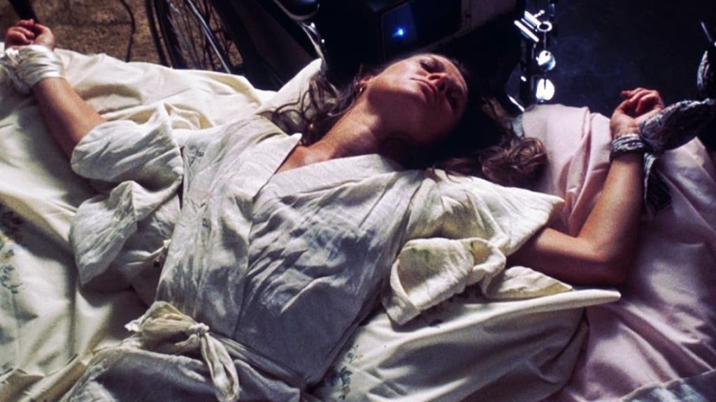 Kvinne kledd i hvitt ligger i en seng, armene er bundet fast.